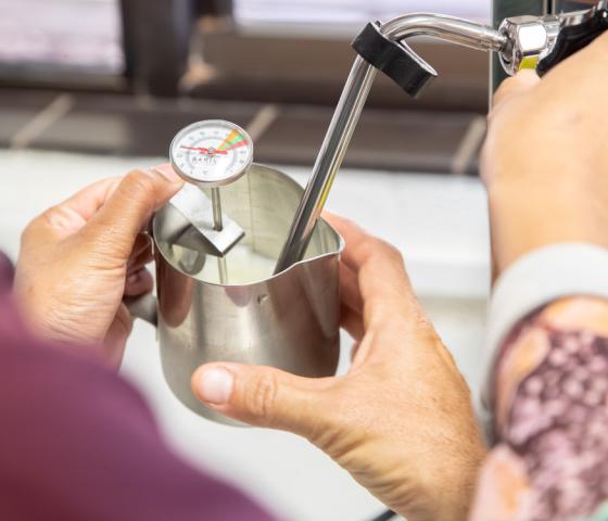 Close-up of a student monitoring milk temperature in a stainless-steel jug while developing barista coffee-making skills.